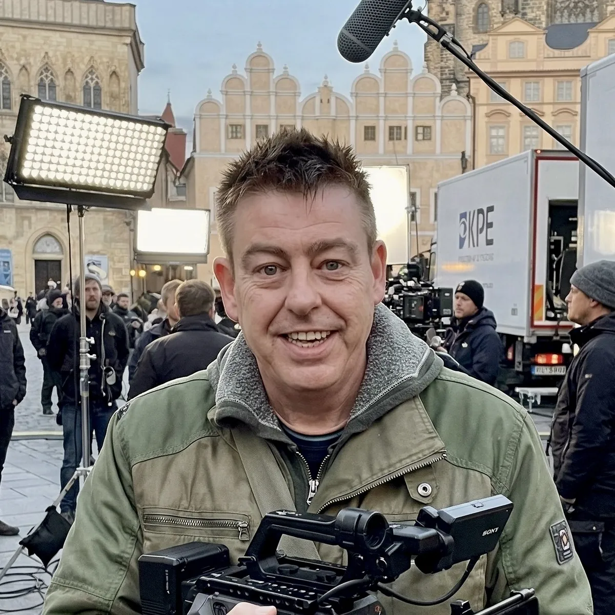 Smiling director holding a camera on a film set in a historic city square, with lighting rigs, a boom microphone and crew behind him