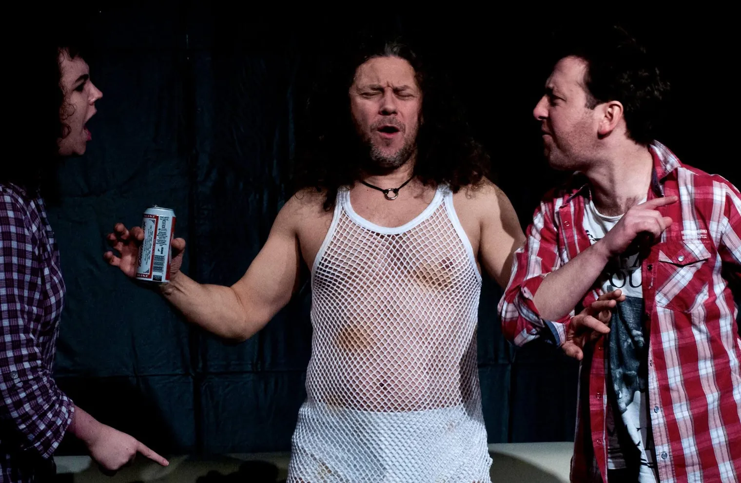Three actors in a heated stage scene: a long-haired man in a string vest holding a beer can is confronted by two others pointing at him